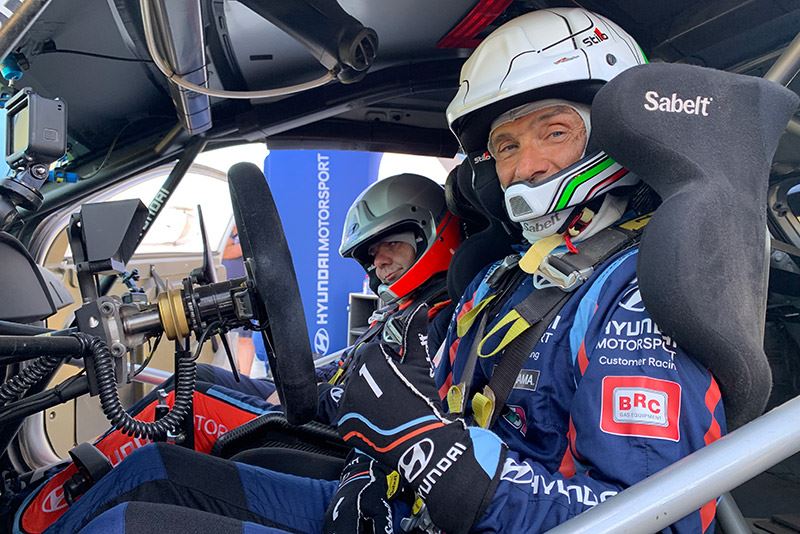 Gabriele Tarquini op de passagiersstoel naast Andreas Mikkelsen, achter het stuur van een rallyauto Hyundai i20 WRC.